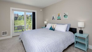 A bedroom with a large bed, two bedside tables with lamps, a sliding glass door to a balcony, and artwork on the wall. - Gorgeous Vacation Chalet: Hot Tub, Wood Stove (Big Sky)