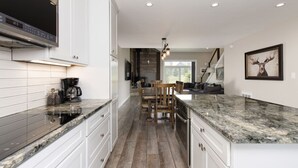 A modern kitchen with white cabinetry, granite countertops, and a built-in oven. There is a dining area with a wooden table and chairs, and a large window providing natural light. - Gorgeous Vacation Chalet: Hot Tub, Wood Stove (Big Sky)