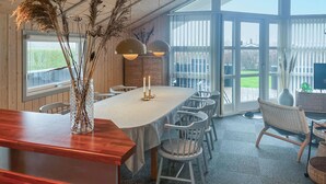En matplats med ett långt bord, vita stolar, en bardisk i trä och ett stort fönster med utsikt över en inhägnad trädgård.