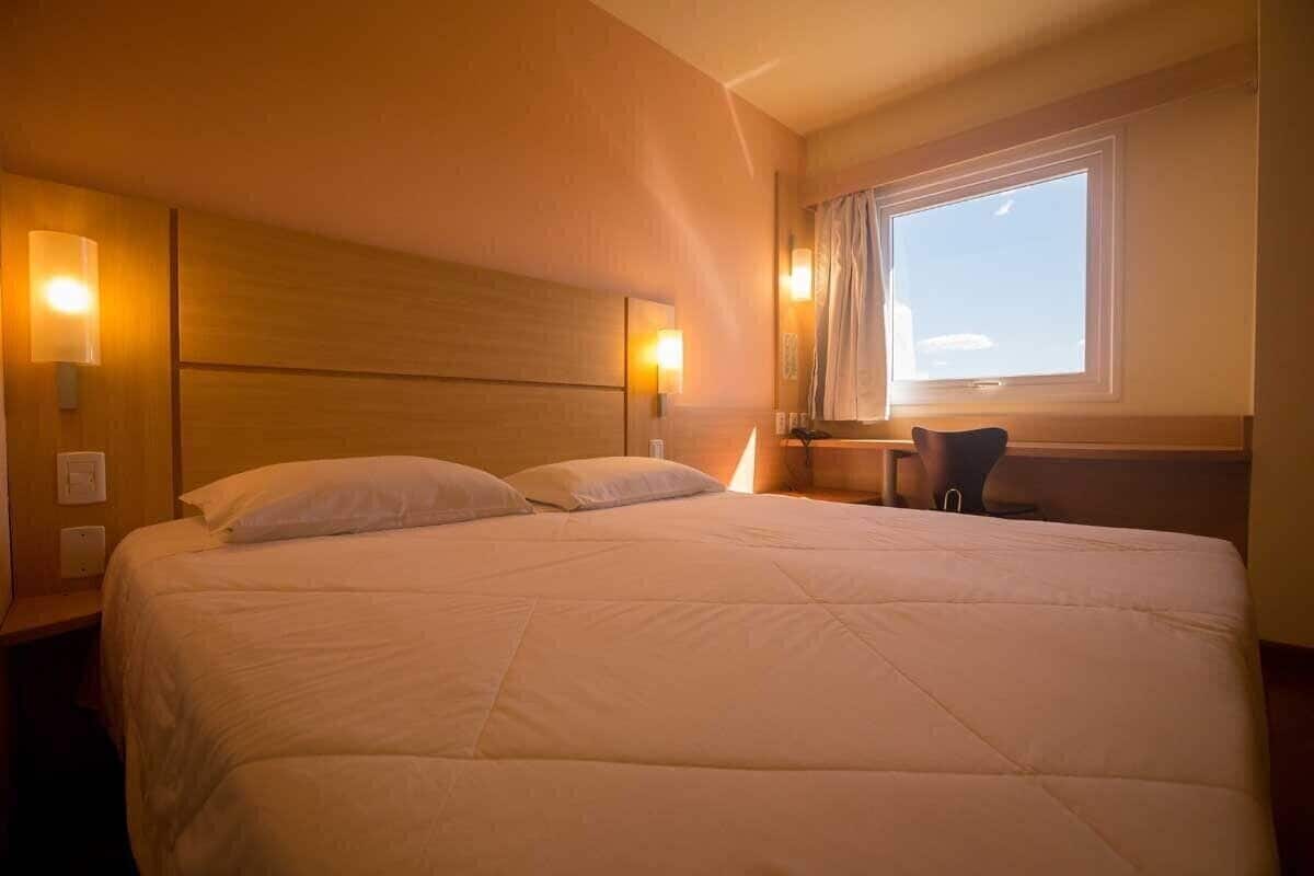 Una habitación de hotel con una cama grande, lámparas de noche, una mesita y un ventanal con cortinas.