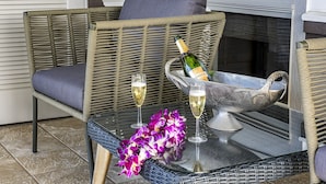 A wicker chair with a cushion, a glass table with a bottle of champagne and two glasses, and purple flowers. - 🏖️Peaceful | Concierge Included | Mauna Lani (Kamuela)