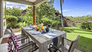 An outdoor dining area with a wooden table set for four, surrounded by chairs, with a view of a lush garden and a clear blue sky. - 🏖️Peaceful | Concierge Included | Mauna Lani (Kamuela)