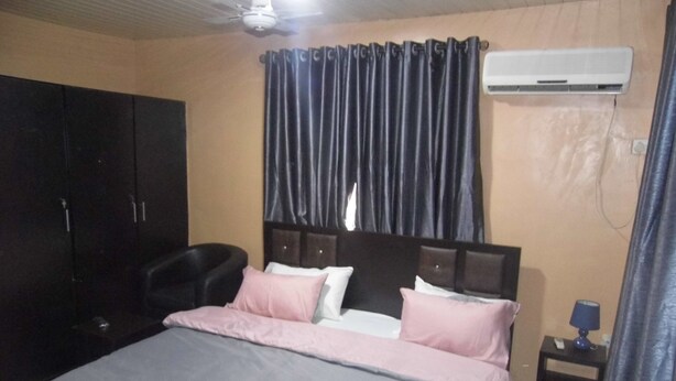 A bedroom with a bed, a wardrobe, a chair, an air conditioning unit, and a nightstand with a lamp.