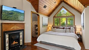 Une chambre avec une cheminée, un grand lit, une télévision et une fenêtre donnant sur les arbres.