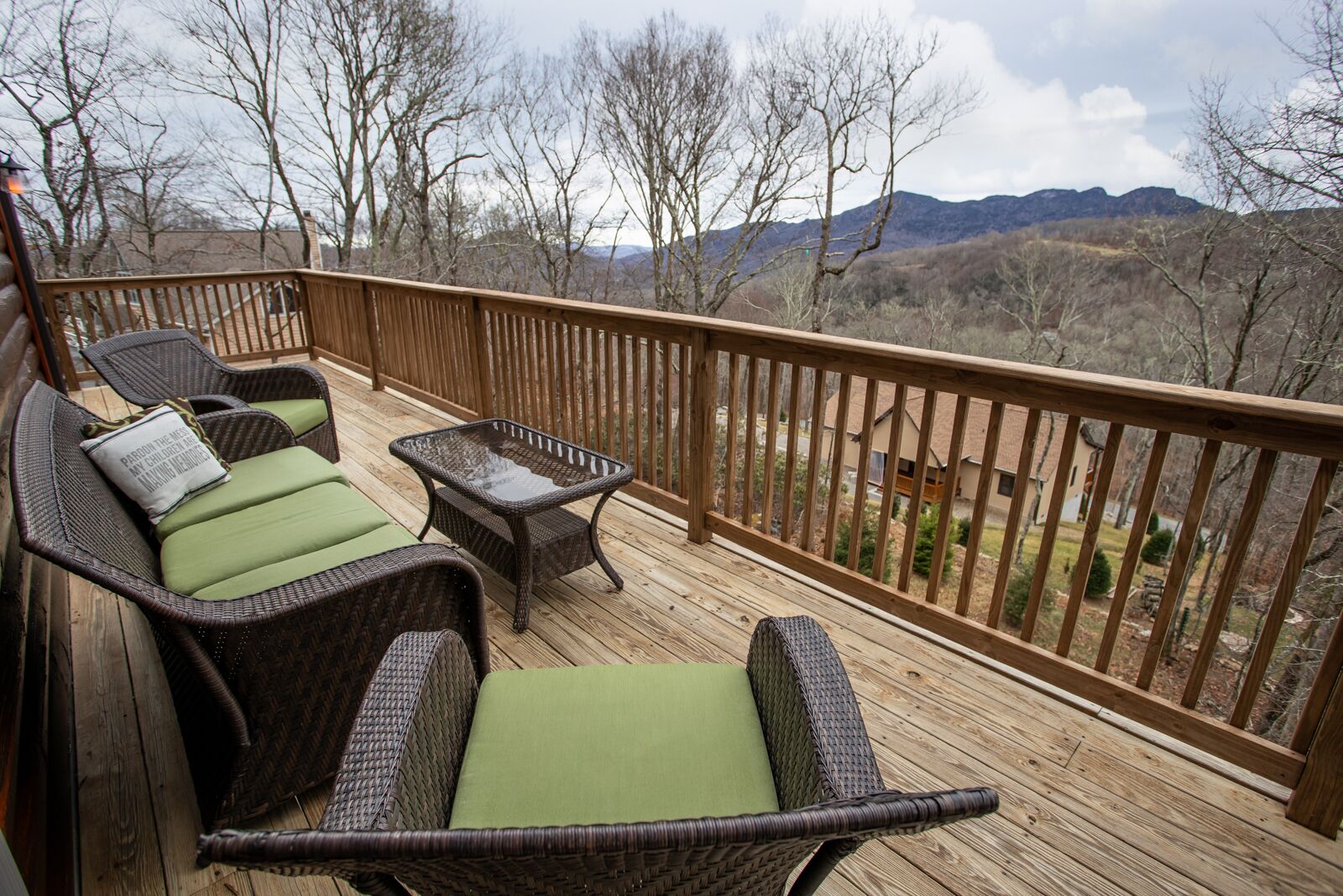A wooden deck with wicker furniture and a glass table, overlooking a mountainous landscape.