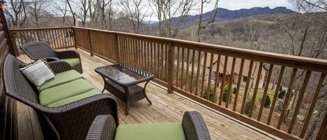 A wooden deck with wicker furniture and a glass table, overlooking a mountainous landscape.