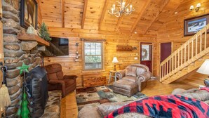 A cozy cabin interior with a stone wall, wooden ceiling, a fireplace, a TV, a staircase, and a large window.