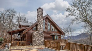 A log cabin with a stone chimney, wooden deck, and a view of mountains.