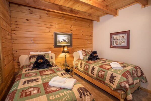 A cabin-style room with two beds, each with a bear-themed quilt, a wooden ceiling, and a framed picture on the wall.