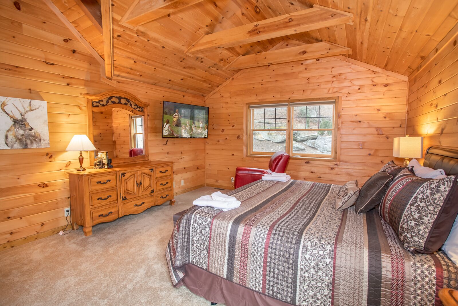 Un dormitorio acogedor tipo cabaña de madera con una cama grande, un aparador, un espejo y una ventana con vistas al exterior.