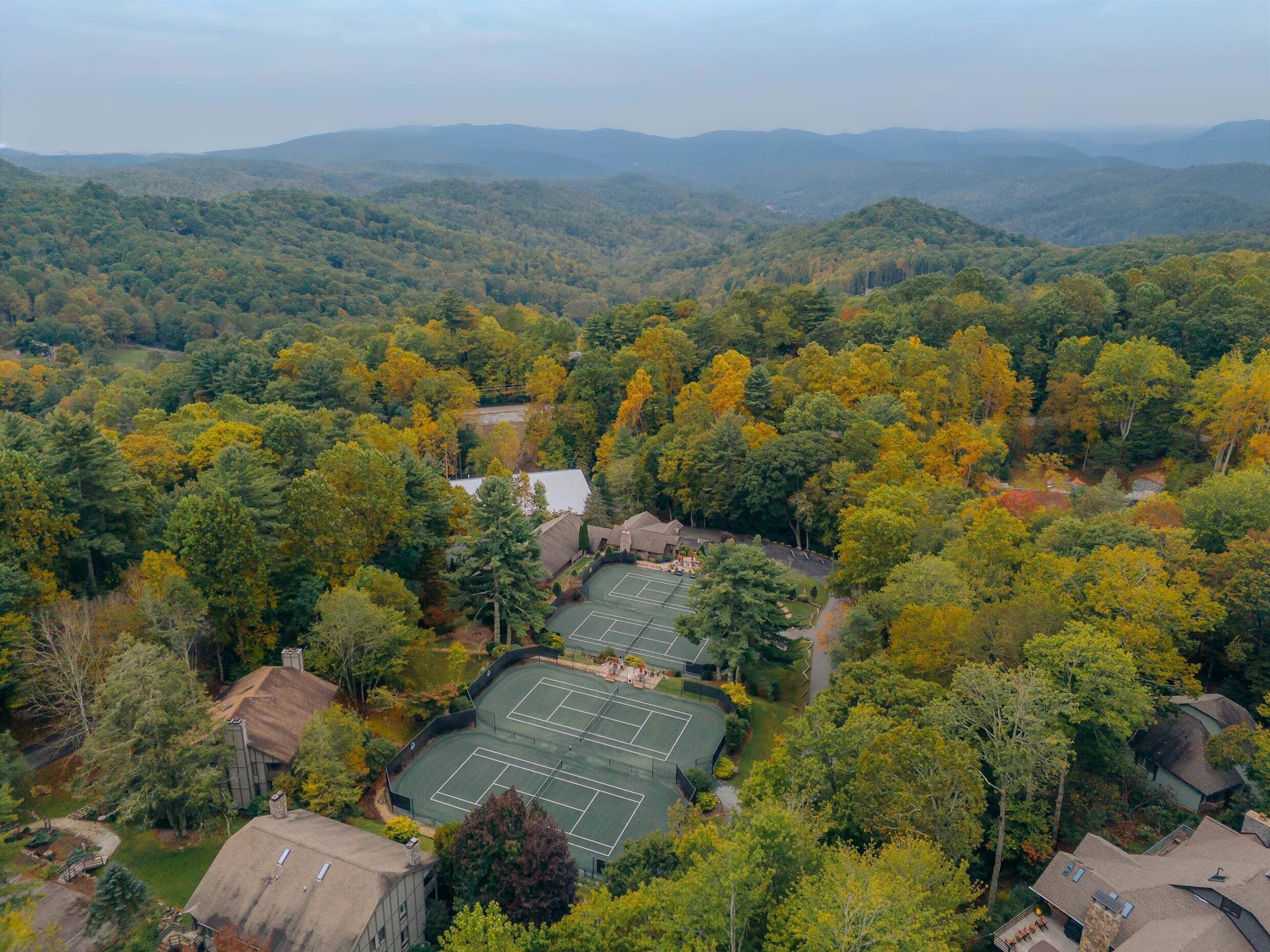 An aerial view of a residential area with multiple tennis courts surrounded by dense forest and hills in the background.