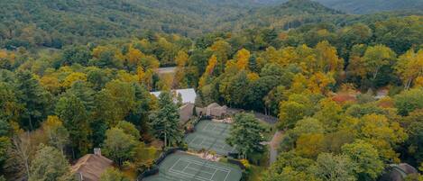 An aerial view of a residential area with multiple tennis courts surrounded by dense forest and hills in the background.