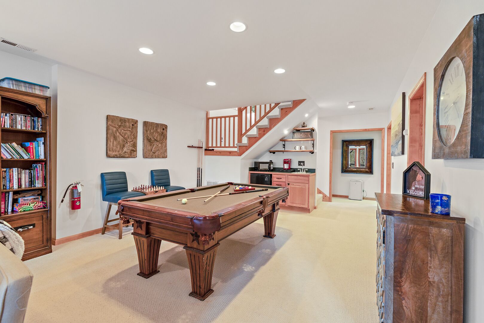 A spacious living room with a pool table, a bookshelf, and a staircase leading to a loft area.