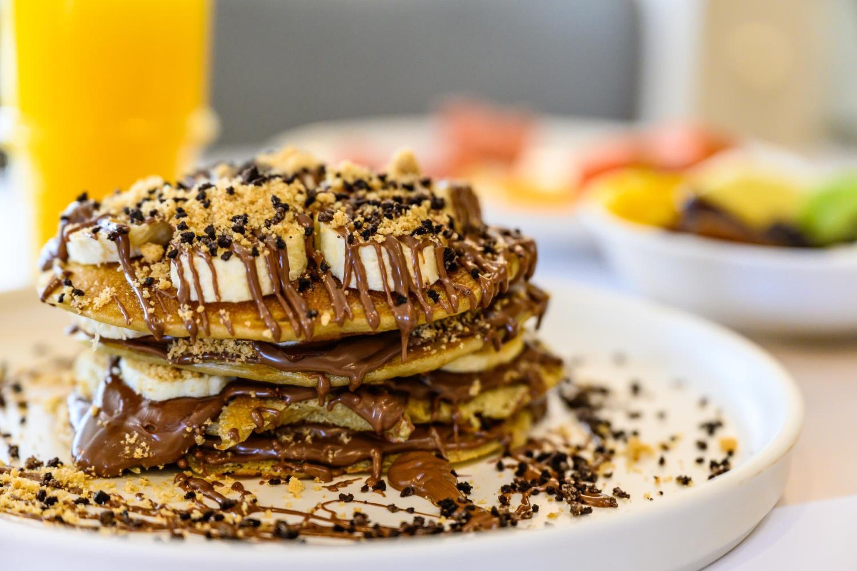 A stack of pancakes with chocolate sauce and crumbled cookies on a white plate.