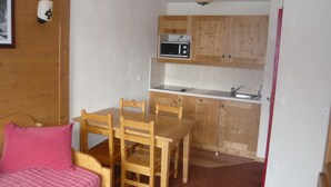 A compact kitchen with wooden cabinets, a microwave, and a sink. There is a dining table with chairs and a bed with a pink pillow. - Apartment in 4 * residence, Les Alpages de Val Cenis (Lanslevillard)
