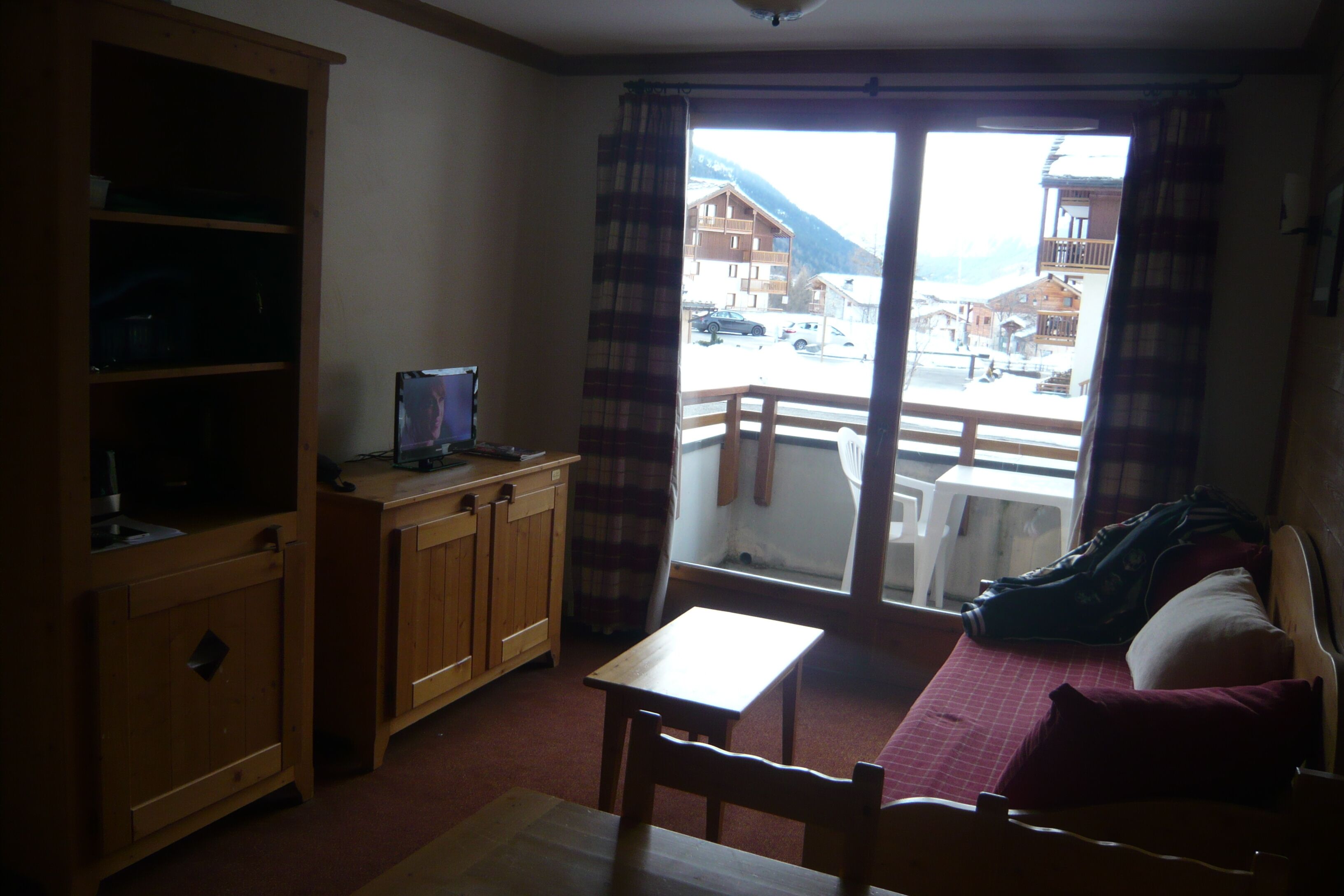 Une pièce avec un meuble en bois, une table, un lit recouvert d’une couverture rouge, et une vue sur une montagne et des bâtiments, visible par une fenêtre.