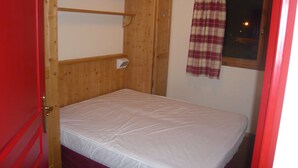 A single bed in a room with a wooden headboard, a window with a plaid curtain, and a red door. - Apartment in 4 * residence, Les Alpages de Val Cenis (Lanslevillard)