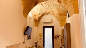 A narrow hallway with a stone archway, a dining area with a table and chairs, a mounted television, and a kitchen area with cabinets and a stove. - Dimora Giulietta Standard Apartment (Lecce)