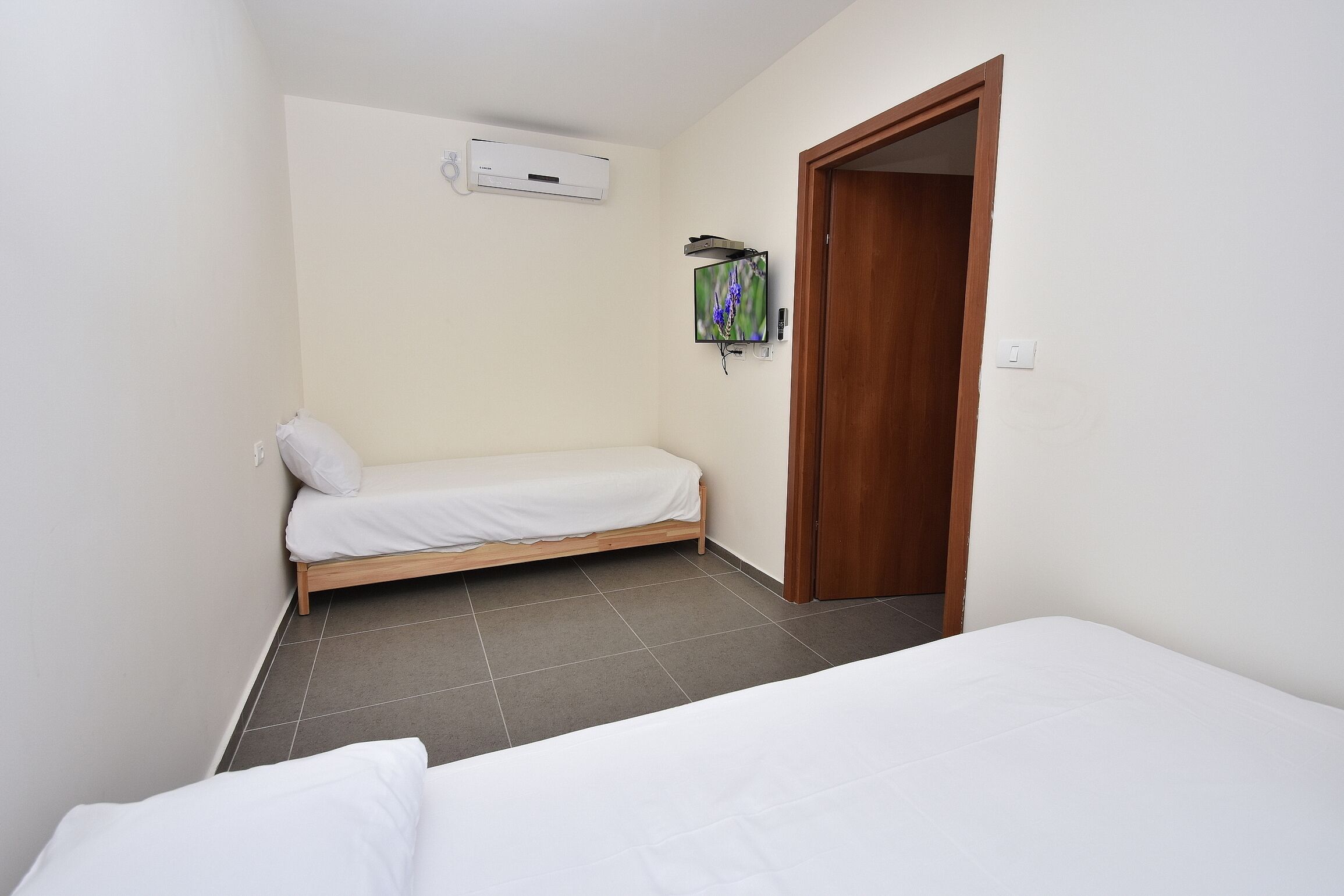 A small, clean room with a single bed, a wooden door, and a mounted TV.