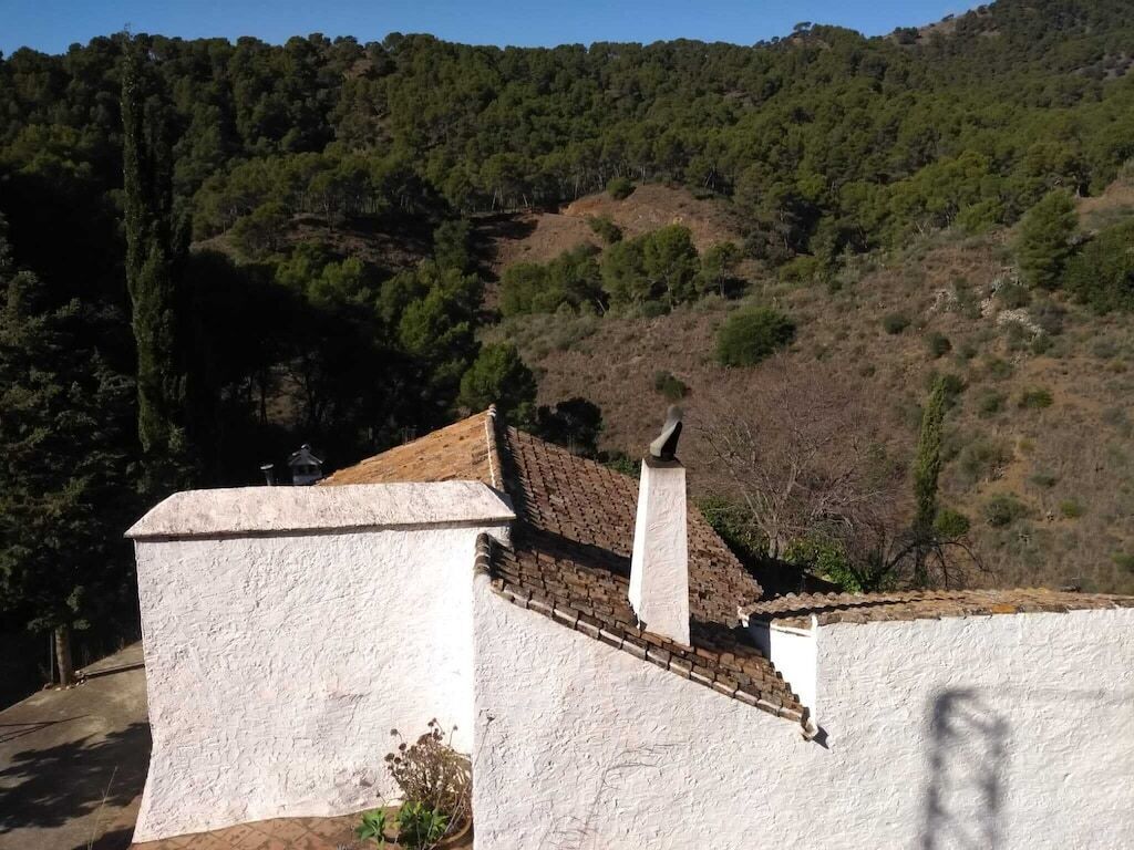 A building with a white exterior and a brick roof, surrounded by a hilly landscape with dense greenery.