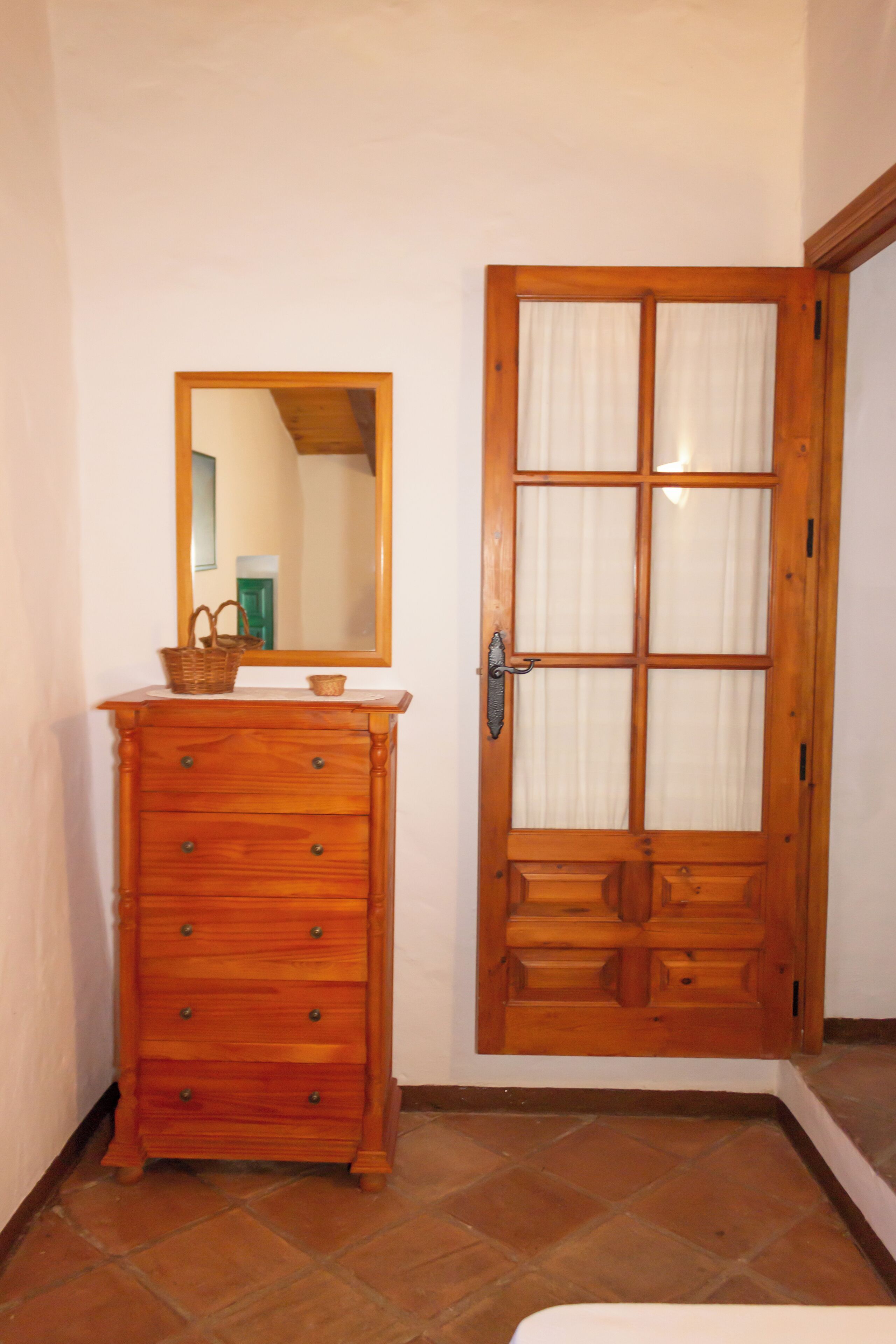 Une commode en bois avec des tiroirs et un miroir, une porte en bois avec des panneaux de verre, et un sol carrelé.