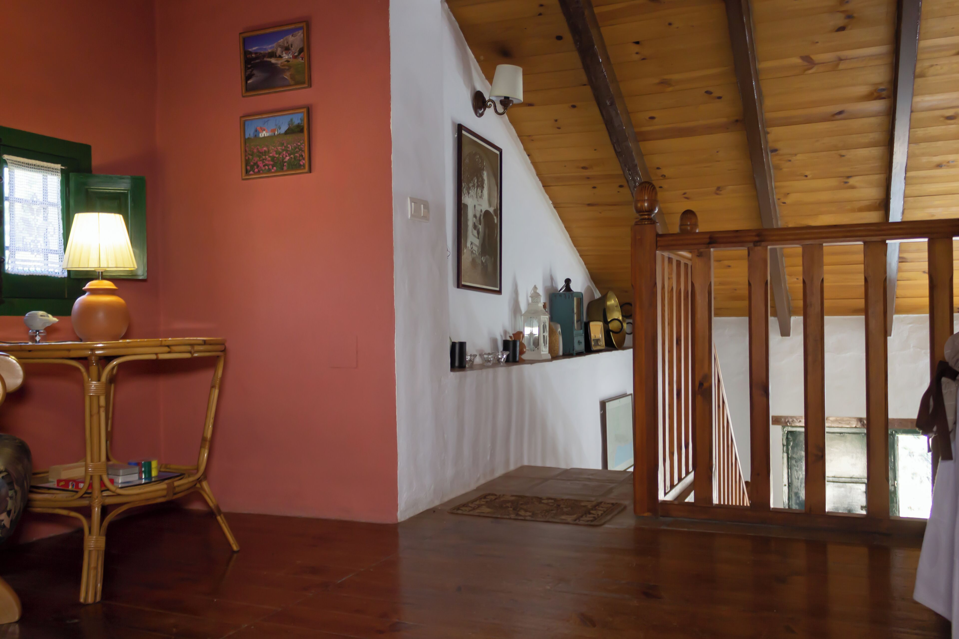 A room with a wooden ceiling, a green-framed window, a wicker table with a lamp, and a wall-mounted shelf with various items.
