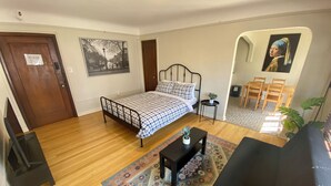 A bedroom with a bed, a small table with a plant, a rug, and a painting on the wall.