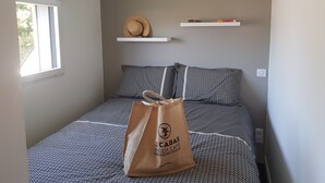 Un dormitorio con una cama estampada, una ventana, un sombrero en un estante y una bolsa de café sobre la cama.