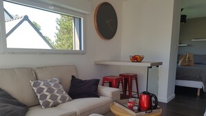 Una sala moderna con sofá, reposapiés y una mesa de centro con una tetera roja. Se ve un estante pequeño con un cuenco de fruta y una bolsa de compras. Al fondo, una cama con edredón estampado es visible.