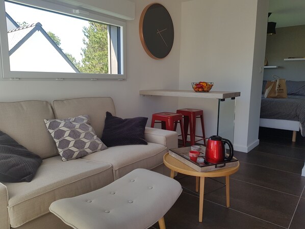 Una sala moderna con sofá, reposapiés y una mesa de centro con una tetera roja. Se ve un estante pequeño con un cuenco de fruta y una bolsa de compras. Al fondo, una cama con edredón estampado es visible.