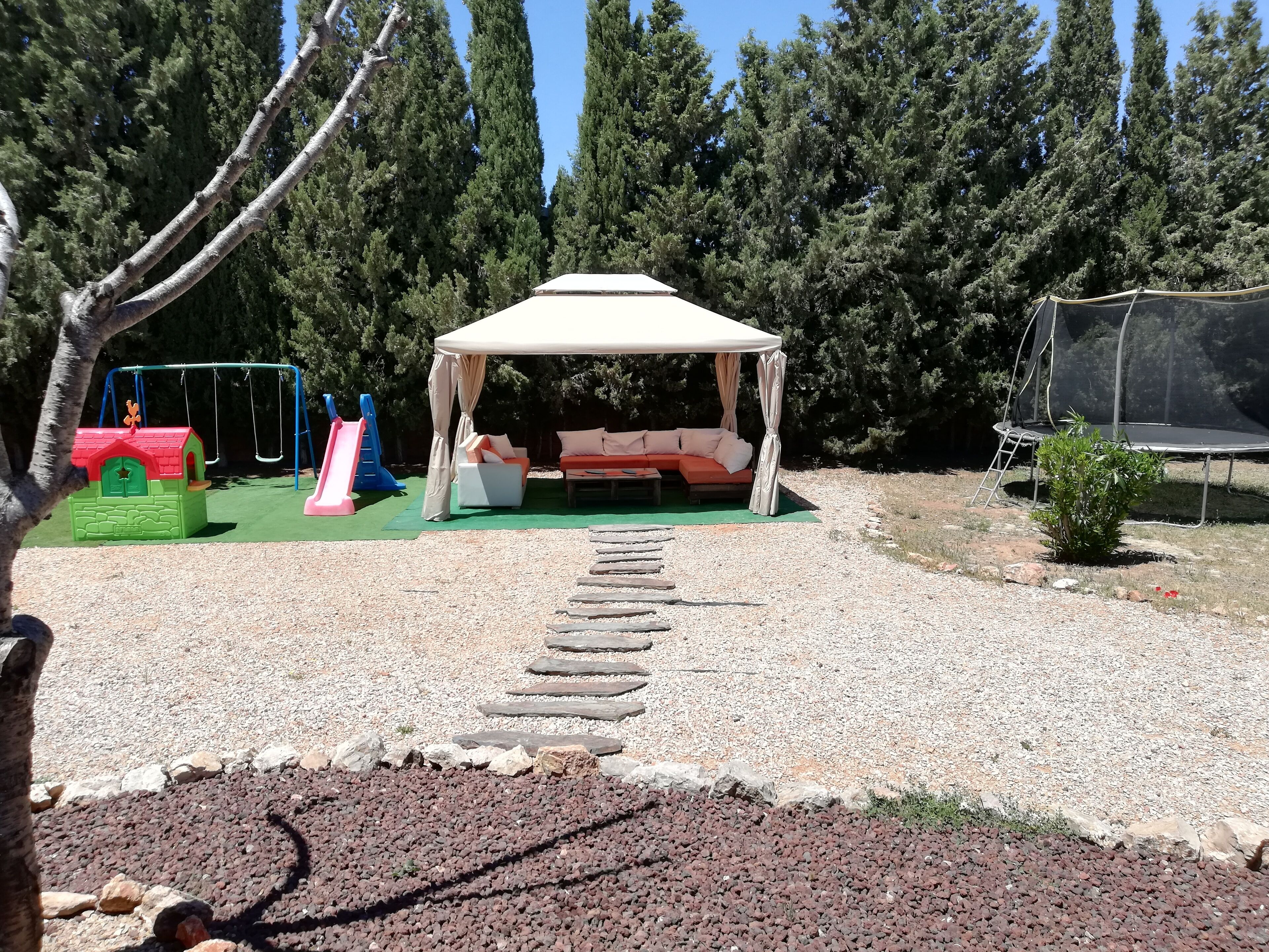 Un espace extérieur aménagé, abrité et doté de sièges, accessible par un chemin de pierre, et entouré d’arbres et d’une aire de jeux.