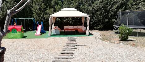 Un espace extérieur aménagé, abrité et doté de sièges, accessible par un chemin de pierre, et entouré d’arbres et d’une aire de jeux.