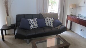 A living room with a black leather sofa, two patterned cushions, a glass coffee table, a small wooden side table, a vase with branches, a lamp, and a framed picture on the wall.