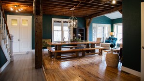 Une pièce spacieuse avec un plancher en bois, une grande table en bois entourée de bancs, un luminaire suspendu et une vue sur l’extérieur par une porte.