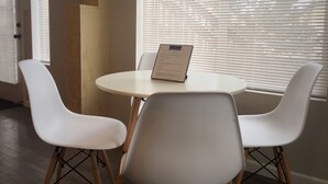 Een kleine, ronde tafel met een menukaart, omringd door twee witte stoelen met houten poten, in een kamer met grote ramen en luiken.