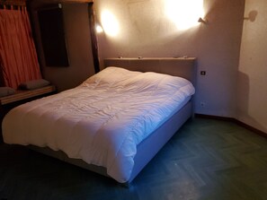 A neatly made bed in a room with a wooden floor, a nightstand with a lamp, and a wall-mounted light fixture.