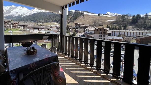 Un balcón con una mesa y sillas, con vistas a un paisaje montañoso que incluye edificios y picos nevados.