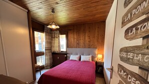 Un dormitorio con techo de madera, una cama con colcha roja, una mesita de noche con lámpara y una ventana con cortinas.