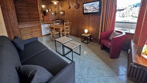 Una sala de estar acogedora con techo de madera, comedor con mesa y sillas de madera, un sillón rojo y un televisor montado en la pared.