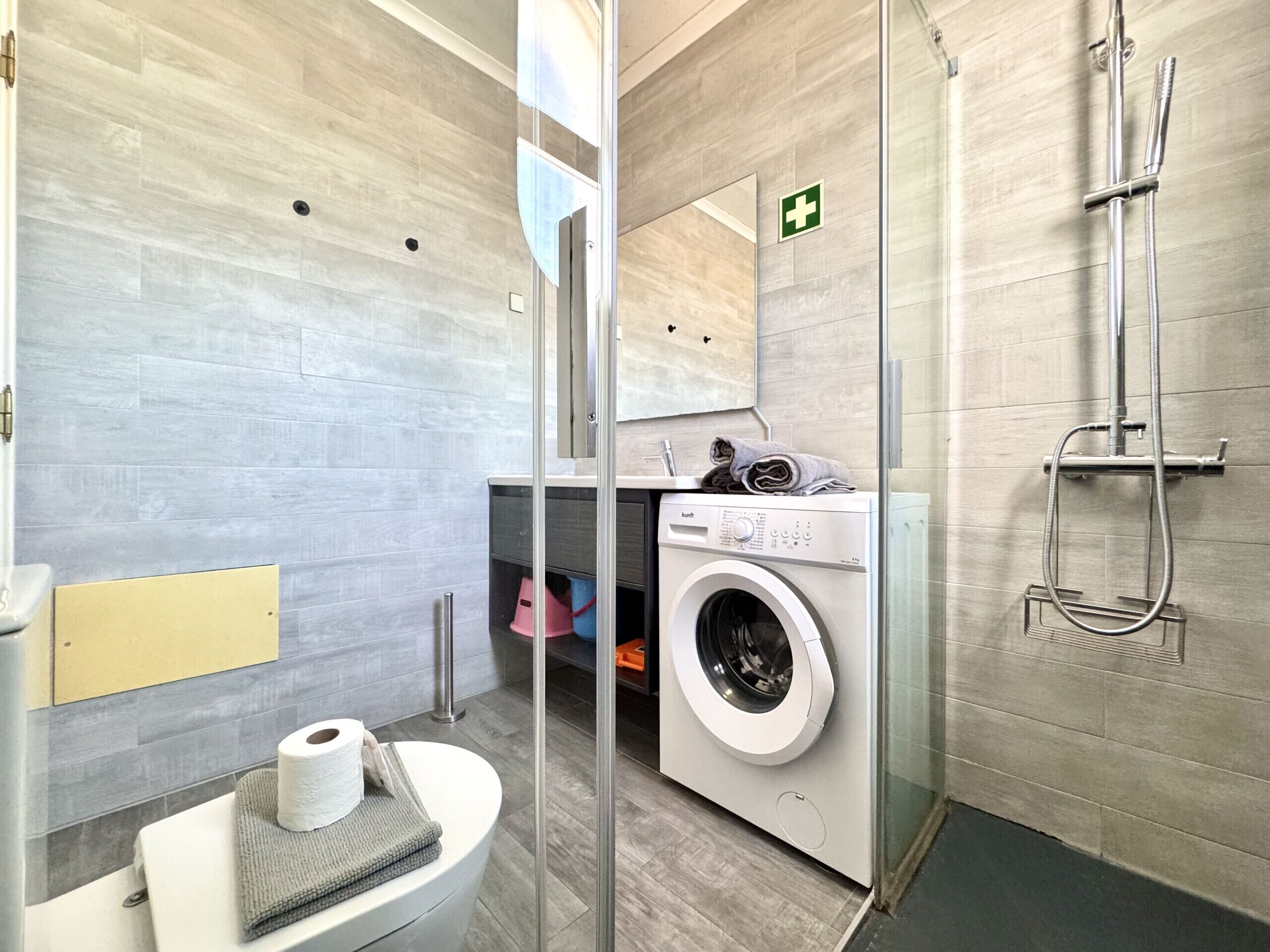 Ein modernes Badezimmer mit Duschkabine aus Glas, integriertem Waschmaschinenanschluss und Toilette mit Handtuch und Toilettenpapier.