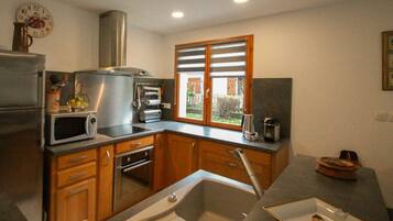A compact kitchen with wooden cabinets, a stainless steel refrigerator, a microwave, an oven, a sink, and a window with blinds.