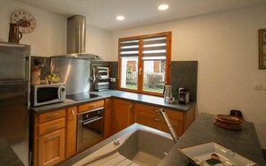 Een kleine keuken met houten kastjes, een roestvrijstalen koelkast, een magnetron, een oven, een spoelbak en een raam met zonwering.
