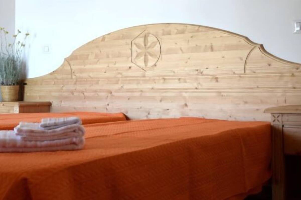 Una habitación con dos camas, cada una con colcha naranja y toallas dobladas con cuidado. Se observa un cabecero de madera con diseño geométrico, una mesita de noche de madera con planta en maceta, e un enchufe de pared.
