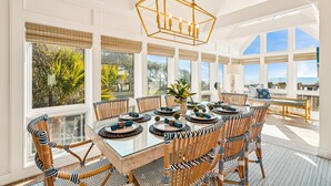 A sunlit dining area with a large table, wicker chairs, and a hanging light fixture.