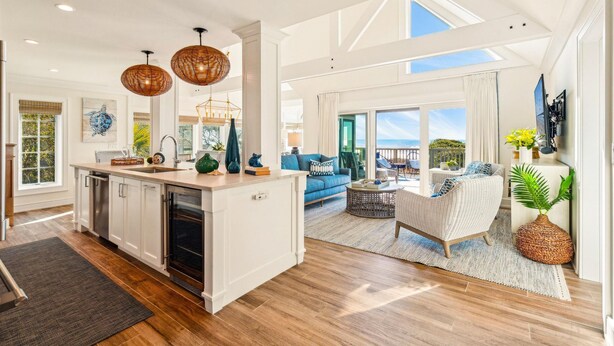 A spacious living area with a large window, a kitchen with white cabinets, and a dining area with a wicker chair.