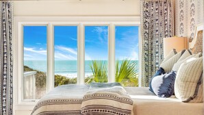 A bedroom with a large window overlooking a beach, a bed with patterned bedding, and a bedside lamp.