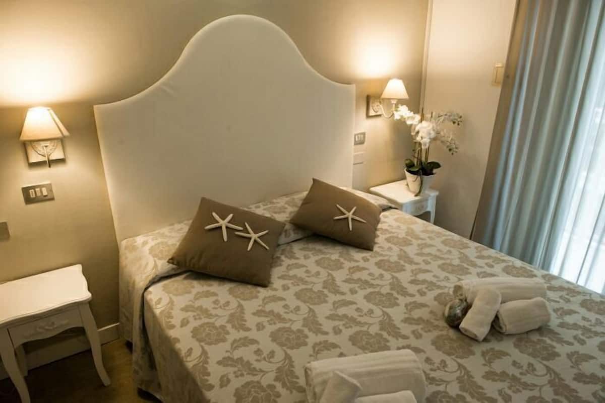 Een net opgemaakt bed met bloemenbedrukte lakens, twee kussens met sterrenvismotieven, een nachtkastje met een lamp en een vaas met witte bloemen.