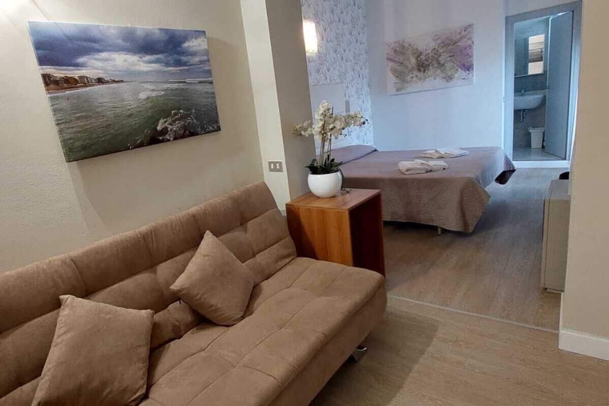 Een moderne woonkamer met een beige bank, een schilderij van een strandgezicht, en een tafeltje met een witte vaas en bloemen.