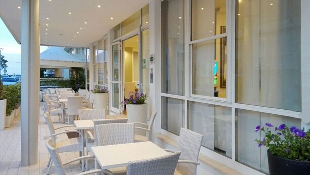 An outdoor seating area with wicker chairs and tables, large windows, and potted plants. - Hotel Ermeti B&B and Brunch (Riccione)