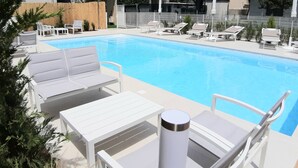 A pool area with lounge chairs and a table, surrounded by a fence and greenery. - Hotel Ermeti B&B and Brunch (Riccione)